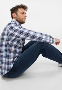 Blau-weiß-rotes kariertes Hemd; kombiniert mit dunkelblauen Jeans und weißen Sneakers; sitzende Pose vor einem neutralen Hintergrund.