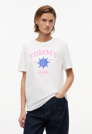 Νεαρό άτομο με κοντά μαλλιά που φοράει λευκό μπλουζάκι "Tommy Jeans" με μπλε γραφικό ήλιου και σκουρόχρωμο τζιν παντελόνι, με το χέρι στην τσέπη.