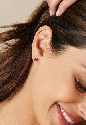Runde Steckerohrringe mit einem dunkelblauen Edelstein, der in einem kleinen metallischen Verschluss gefasst ist, der am Ohr eines Models vor einem neutralen Hintergrund präsentiert wird.