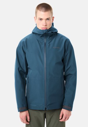 Junger Mann, der eine blaue wasserdichte Kapuzenjacke trägt, die hochgeschlossen ist, steht vor einem hellgrauen Hintergrund mit entspannt an den Seiten hängenden Händen.