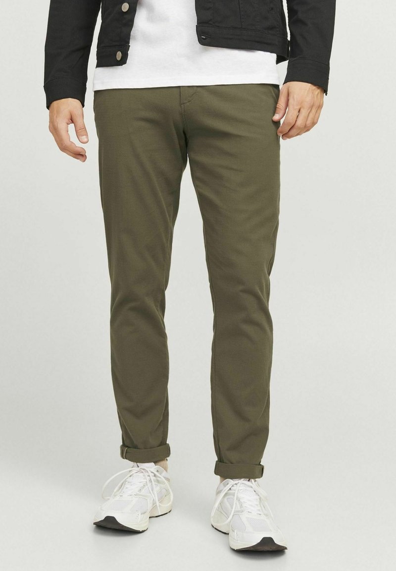 Pantaloni verzi măslinii cu un croi slim, realizați dintr-un material ușor. Au margini răsucite, buzunare frontale și sunt asortați cu pantofi sport albi.