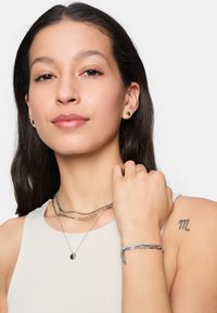 Collane impilabili argentate con forme varie, accentuate da un bracciale elegante. La modella indossa orecchini a bottone rotondi e un top aderente.