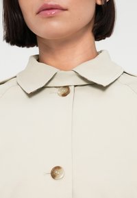 Μπεζ παλτό με απαλή υφή, που διαθέτει μεγάλο γιακά και λαμπερό, στρογγυλό κουμπί στη λαιμόκοψη. Καθαρός, μινιμαλιστικός σχεδιασμός.