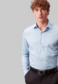 Jeune homme aux cheveux roux bouclés portant une chemise bleu clair à boutons et un pantalon foncé avec une ceinture noire, debout devant un fond gris.