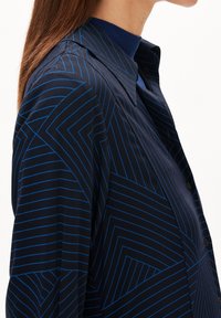 Kvinna som bär en marinblå skjorta med geometriska blå linjemönster och en mörkblå polokrage under, visad från sidan.