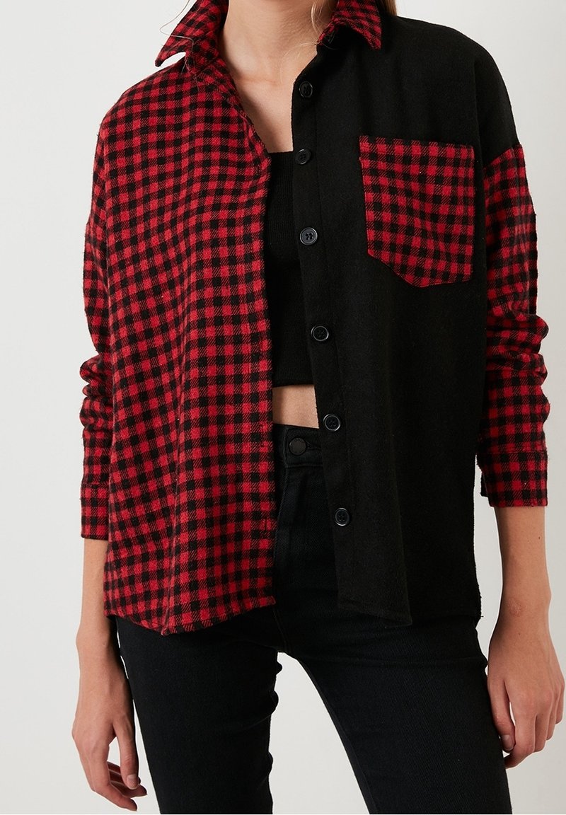 Camisa de franela a cuadros en negro y rojo con cierre de botón, mangas largas y un bolsillo en el pecho. La tela parece suave y texturizada.