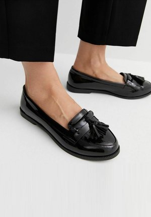 Pieds portant des mocassins à pampilles en cuir verni noir et un pantalon noir raccourci sur un fond clair uni.