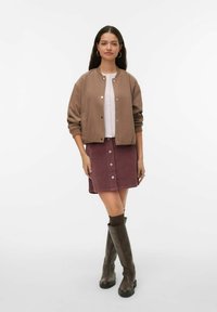 Veste aviateur marron, chemise rose clair avec boutons, jupe en velours côtelé bordeaux avec boutons sur le devant, et bottes en cuir marron montant jusqu'aux genoux.