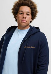 Marineblauwe zip-up hoodie met geborduurde tekst "Originals Studio" in goud, gecombineerd met een lichtblauw T-shirt. Heeft een capuchon met trekkoord.