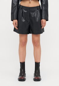 Shorts en cuir synthétique noirs avec une texture lisse, un design plissé et des passants de ceinture. Assortis à des bottes noires brillantes jusqu'aux genoux avec des accents rouges.
