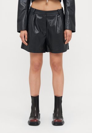 Shorts en cuir synthétique noirs avec une texture lisse, un design plissé et des passants de ceinture. Assortis à des bottes noires brillantes jusqu'aux genoux avec des accents rouges.