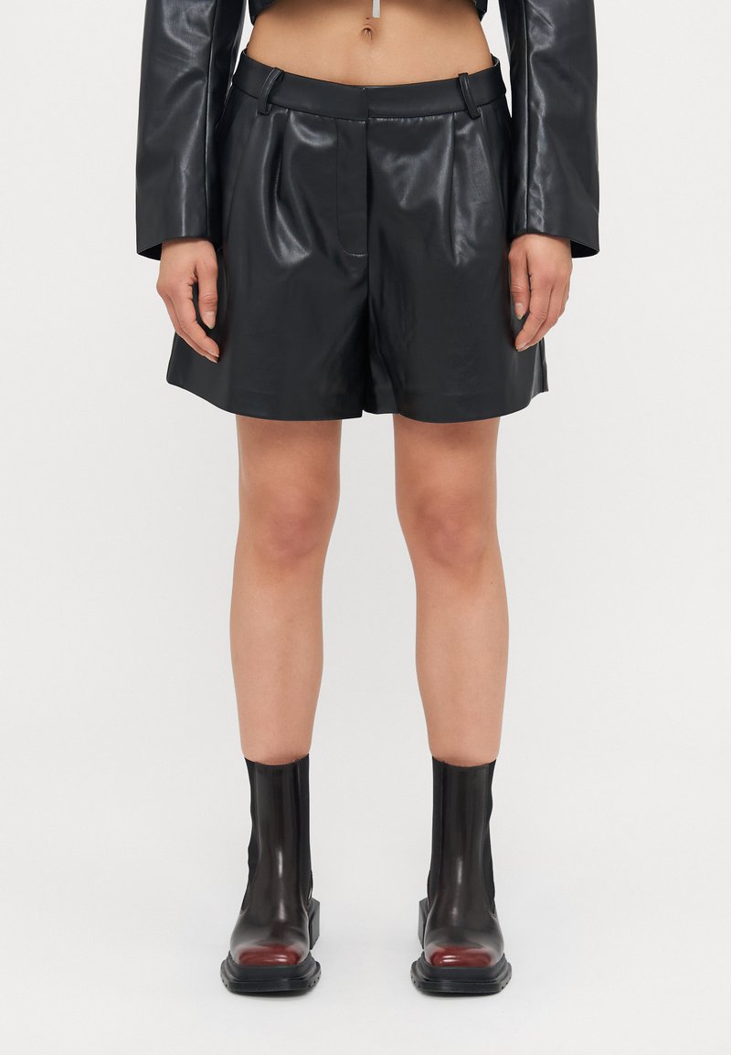 Shorts en cuir synthétique noirs avec une texture lisse, un design plissé et des passants de ceinture. Assortis à des bottes noires brillantes jusqu'aux genoux avec des accents rouges.