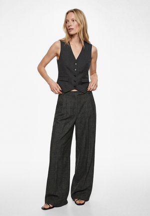 Vrouw met een zwart vest met knopen en wijde donkergrijze geruite broek, staand met handen bij de taille, kijkend naar links.