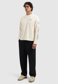 Cream long-sleeve shirt with black graphic text along one sleeve, paired with black pants and light-colored shoes on a neutral background.