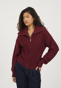 Burgundowa dzianinowa sweter z wysokim kołnierzem, zapięciem na zamek, prążkowaną fakturą i szerokimi rękawami. Połączony z ciemnoniebieskimi spodniami.