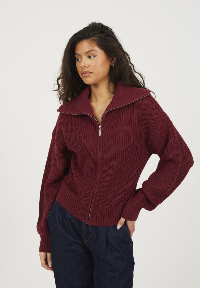 Burgundowa dzianinowa sweter z wysokim kołnierzem, zapięciem na zamek, prążkowaną fakturą i szerokimi rękawami. Połączony z ciemnoniebieskimi spodniami.