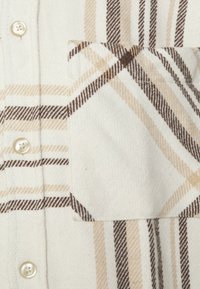 Camicia di flanella a quadri in crema, marrone e beige, con una tasca sul petto, chiusura a bottoni e una texture morbida. Motivo a righe su tutta la superficie.