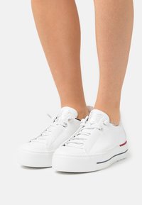 Weiße Ledersneaker mit strukturierten Zungen, flachen Schnürsenkeln und Plateausohlen, die blaue und rote Streifen an der Seite aufweisen.