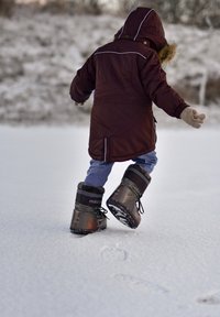 Ruda parka su kailiu apvilktu gobtuvu, šviesiai mėlyni džinsai ir juodi žieminiai batai su pilkais akcentais, einant po sniegu padengtą žemę.
