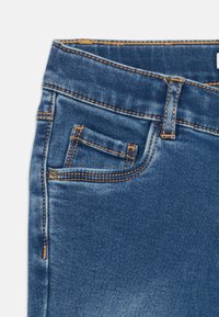 Blue denim jeans featuring a front pocket with orange stitching and a classic five-pocket design. The fabric appears smooth and slightly glossy.