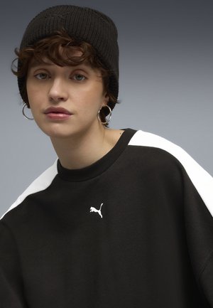 Black sweatshirt with white shoulder accents and a small white Puma logo. Model wears a black knit beanie and hoop earrings. Smooth texture.