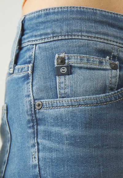 Jeans din denim albastru cu o textură ușor estompată, având un etichetă cu logo pe buzunarul din spate și nituri din metal negru.