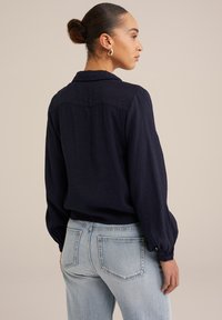 Tumši zilā blūze ar garām, pūkainām piedurknēm, teksturēta auduma, un ar aizmugures joku detalizāciju, apvienota ar gaiši ziliem džinsiem.