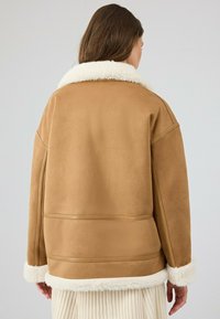 Koffiekleurige suède jas met een romig schapenvachtkraag en -manchetten, met strakke lijnen en een losse pasvorm, die een moderne uitstraling benadrukt.