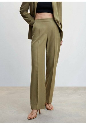 Vrouw draagt olijfgroene op maat gemaakte hooggetailleerde broek gecombineerd met bijpassende blazer, zwart cropped top en beige hakken sandalen, staand op een betonnen vloer.