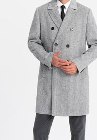 Homme portant un manteau croisé en chevron gris sur une chemise blanche et une cravate noire, les mains jointes près de la taille.