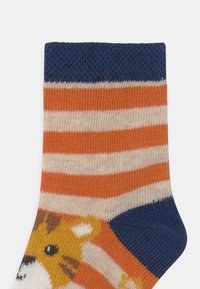 Stribet sok med orange, creme og marineblå farver. Har et mønster med et løveansigt og ribbet kant. Fremstillet af et blødt, strikket materiale.