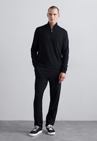 Mann, der an einer einfachen Wand steht, trägt einen schwarzen Reißverschluss-Pullover, schwarze Hosen und schwarze Sneakers mit weißen Sohlen und Schnürsenkeln.