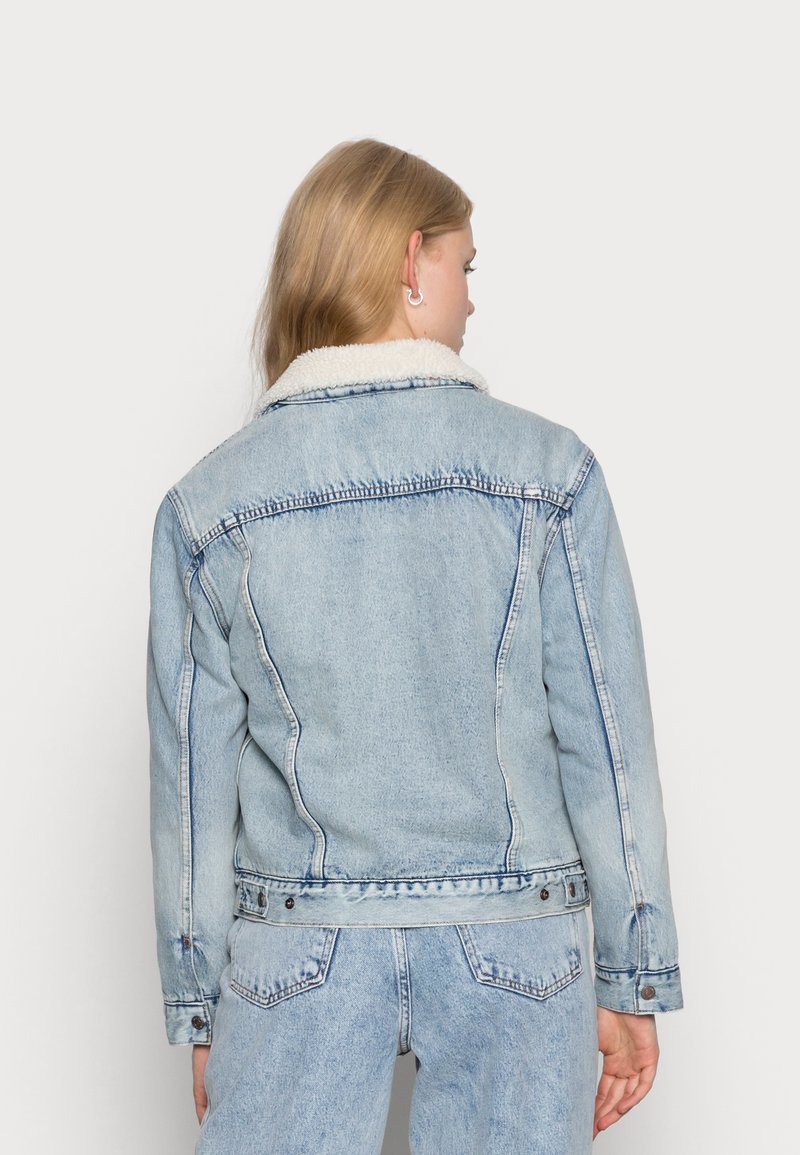 Veste en denim bleu clair avec un col en sherpa, présentant des coutures de poitrine et de dos, des poches latérales et des fermetures à boutons.