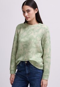 Ljusgrön sweatshirt med ett strukturerat fiskbensmönster, lång ärm och rundhalsringning, tillsammans med mörkblå jeans.