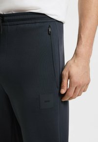 Navy joggerit, joissa on slim fit -leikkaus, vetoketjullinen sivutasku, teksturoitu kangas ja matta logopatchi alaosassa.