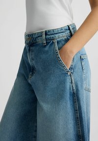 Jeans in denim azzurro chiaro con una vestibilità rilassata; le caratteristiche includono una vita alta, tasche laterali e cuciture distintive lungo le cuciture.