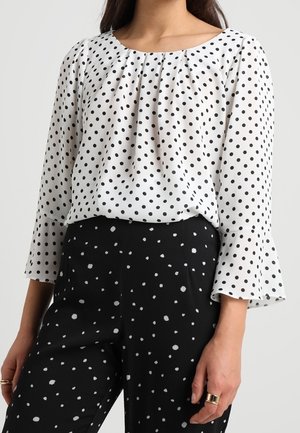 Mujer con blusa blanca de manga larga con lunares negros y pantalones negros con puntos blancos dispersos, mostrando anillos en sus dedos.