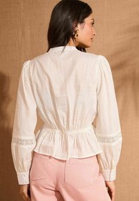 Blusa bianca a trama con maniche lunghe, vita arricciata e dettagli in pizzo sui polsini, abbinata a pantaloni rosa chiaro.