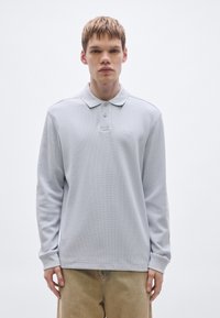 Ανοιχτό γκρι μακρυμάνικο πόλο πουκάμισο με υφή υφάσματος, κούμπωμα με δύο κουμπιά και κεντημένο λογότυπο στο αριστερό στήθος. Άνετη εφαρμογή.