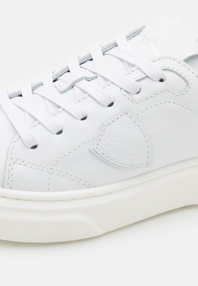 Baskets en cuir blanc avec bout arrondi, lacets plats blancs, détail de logo subtil sur le côté et semelle en caoutchouc texturé blanche.