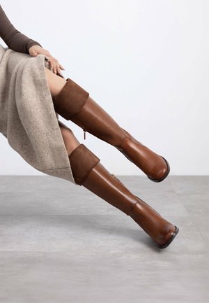 Botas de cuero marrón hasta la rodilla con un puño de ante, que cuentan con una cremallera lateral y una punta redondeada, sobre un fondo neutro.