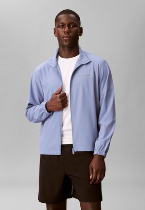 Mand med mørk hud iført en lyseblå lynlåstrøje over en hvid skjorte og sorte shorts, stående mod en grå baggrund.