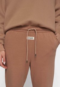 Brune bomulds joggers med elastisk talje, snøre, sidelommer og et lille mærke med teksten "10DAYS." Glat, blød tekstur.