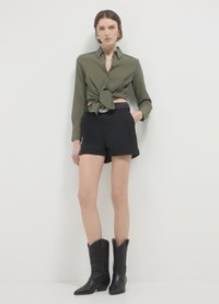 Camicia verde oliva a maniche lunghe con un nodo sul davanti; pantaloni corti neri con orli ripiegati; stivali cowboy neri con design embossato.