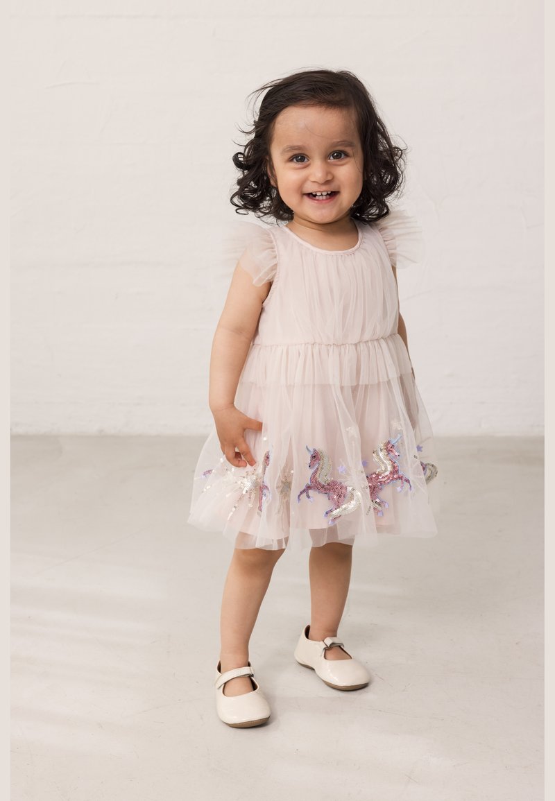 Μικρή κοριτσάκι που χαμογελά, φοράει ένα ανοιχτό ροζ φόρεμα με πούλιες με μονόκερους και λευκά παπούτσια, στέκεται σε έναν απλό φωτεινό δάπεδο και φόντο τοίχου.