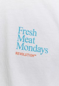 Witte katoenen T-shirt met daarop "Fresh Meat Mondays" in lichtblauw en "REVOLUTION™" in oranje. Zachte textuur met duidelijke, contrasterende tekst.