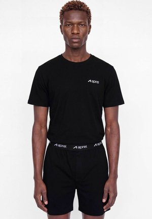 Un mannequin masculin se tient face à l’objectif, vêtu d’un T-shirt noir et d’un short ornés de logos « aprel » blancs, sur un fond blanc uni.