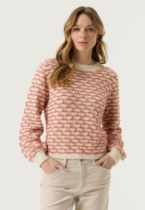 Vrouw met lang golvend haar draagt een gestructureerde roze en crèmekleurige trui en crèmekleurige broek, staat met handen in de zakken tegen een effen achtergrond.