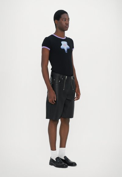 Schwarzes, tailliertes T-Shirt mit lila Akzenten und einem blauen Grafikmotiv, kombiniert mit schwarzen Leder-Shorts mit Reißverschlussdetails und einer glatten Textur.