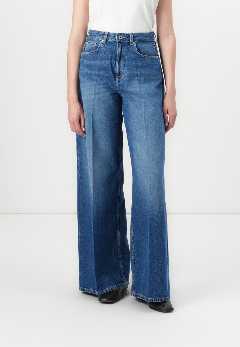 Jeans de denim azul de perna larga com cintura alta, apresentando um fecho de botão, cinco bolsos e costura limpa. Tecido texturizado para durabilidade.